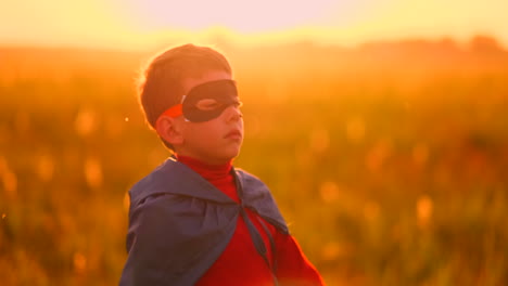 A-child-in-the-costume-of-a-superhero-in-a-red-cloak-runs-across-the-green-lawn-against-the-backdrop-of-a-sunset-toward-the-camera.